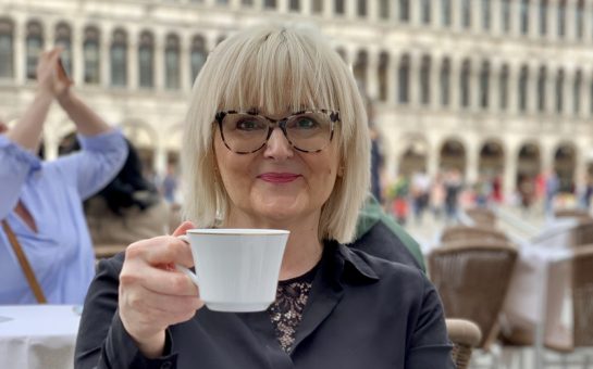 A photo of a blond woman wearing glasses, smiling and holding a tea cup.