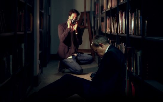 A dark scene showing a man slumped over in a corridor and a person taking a picture of him