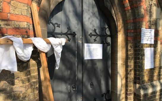 Photo of the front door of a church, closed. On the left of the image there is a wooden cross set up beside the door, with white linen draped over it.
