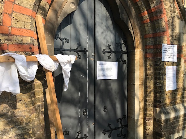Photo of the front door of a church, closed. On the left of the image there is a wooden cross set up beside the door, with white linen draped over it.