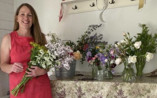 Image shows a white woman in a pink dress holding flowers and standing beside many colourful bouquets.