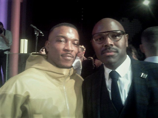 Ashley Walters, a British rapper, in a yellow coat, smiling next to Mr Gee in glasses and a smart tartan suit.