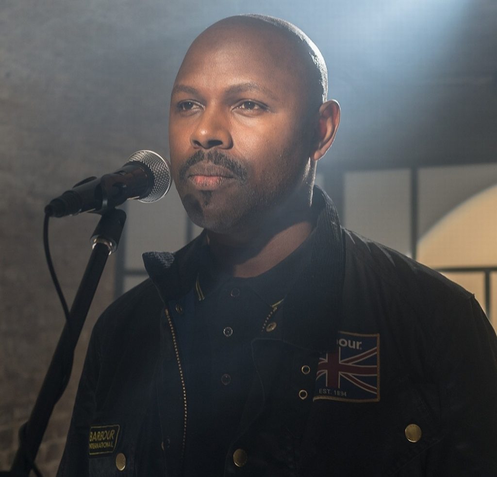 Mr Gee looking stern about to talk into a microphone. There is a spotlight on him. He is bald with a moustache and a goatee.