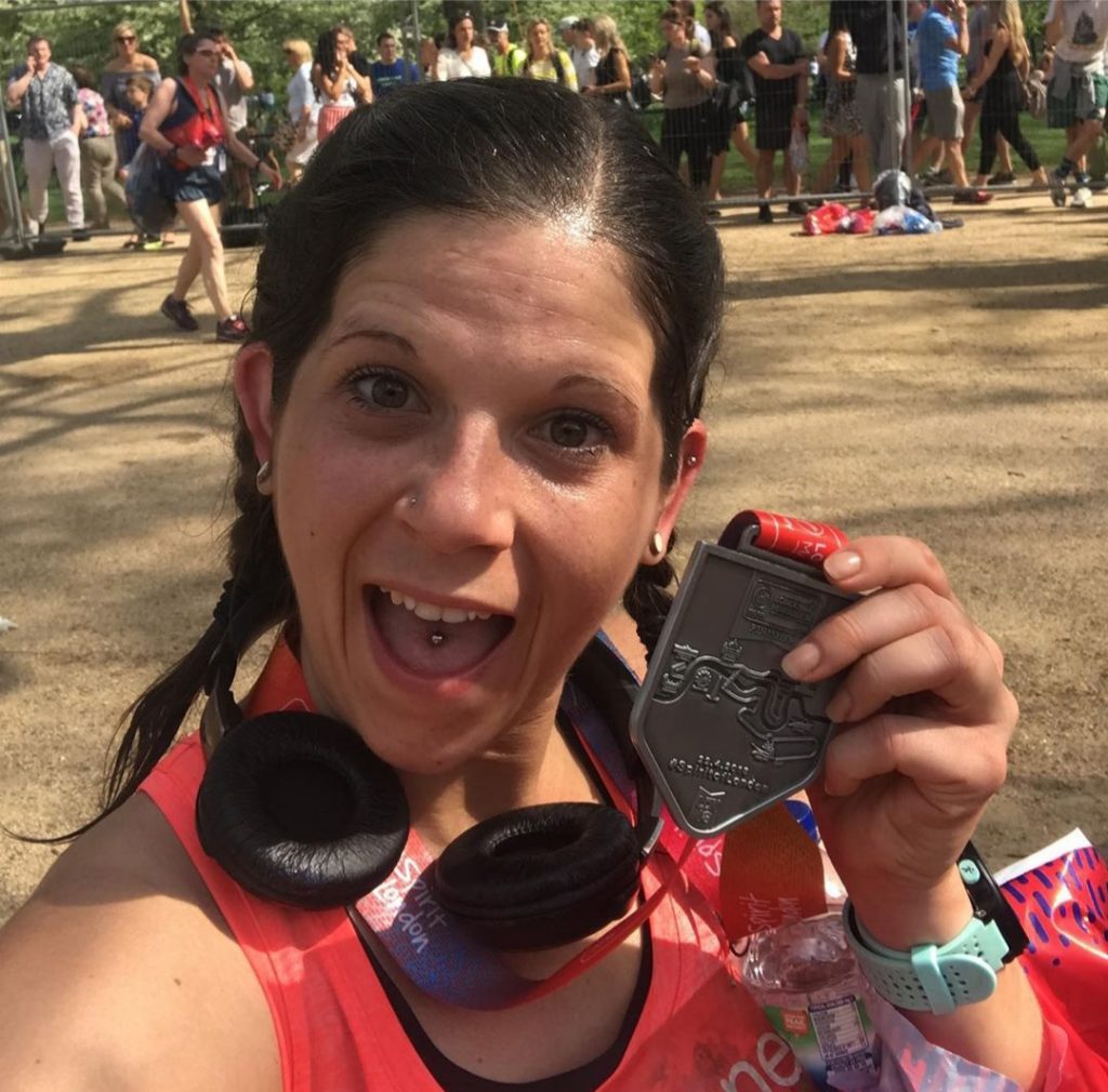 Anna from The Running Channel with a big grin posing with her London Marathon medal. She is wearing a pinky orange sleeveless top, she is wearing a running watching with a light blue strap and she has black headphone around her neck. Her dark brown hair is in plaits resting on her shoulders.