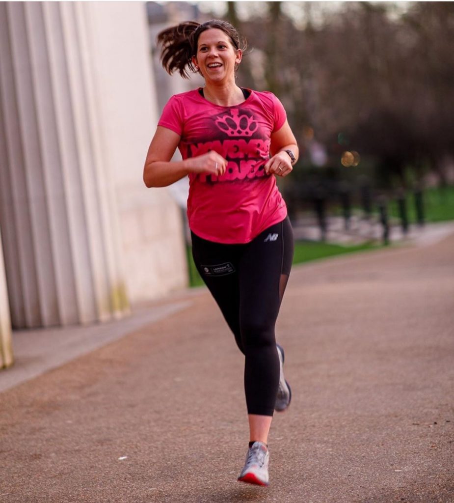 A portrait photo of Anna from The Running Channel running wearing black lycra running leggings and a pink top. She is smiling and her long dark brown pony tail is swishing in the wind.