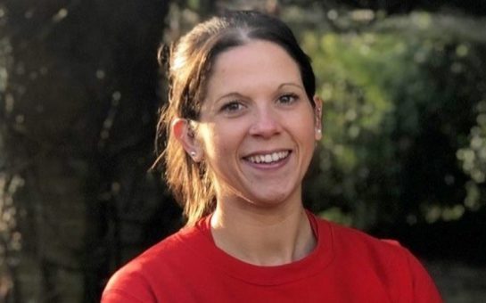 Anna from The Running Channel in a red t-shirt smiling naturally at the camera. She is outside in front of some trees and her dark brown hair is tied back.