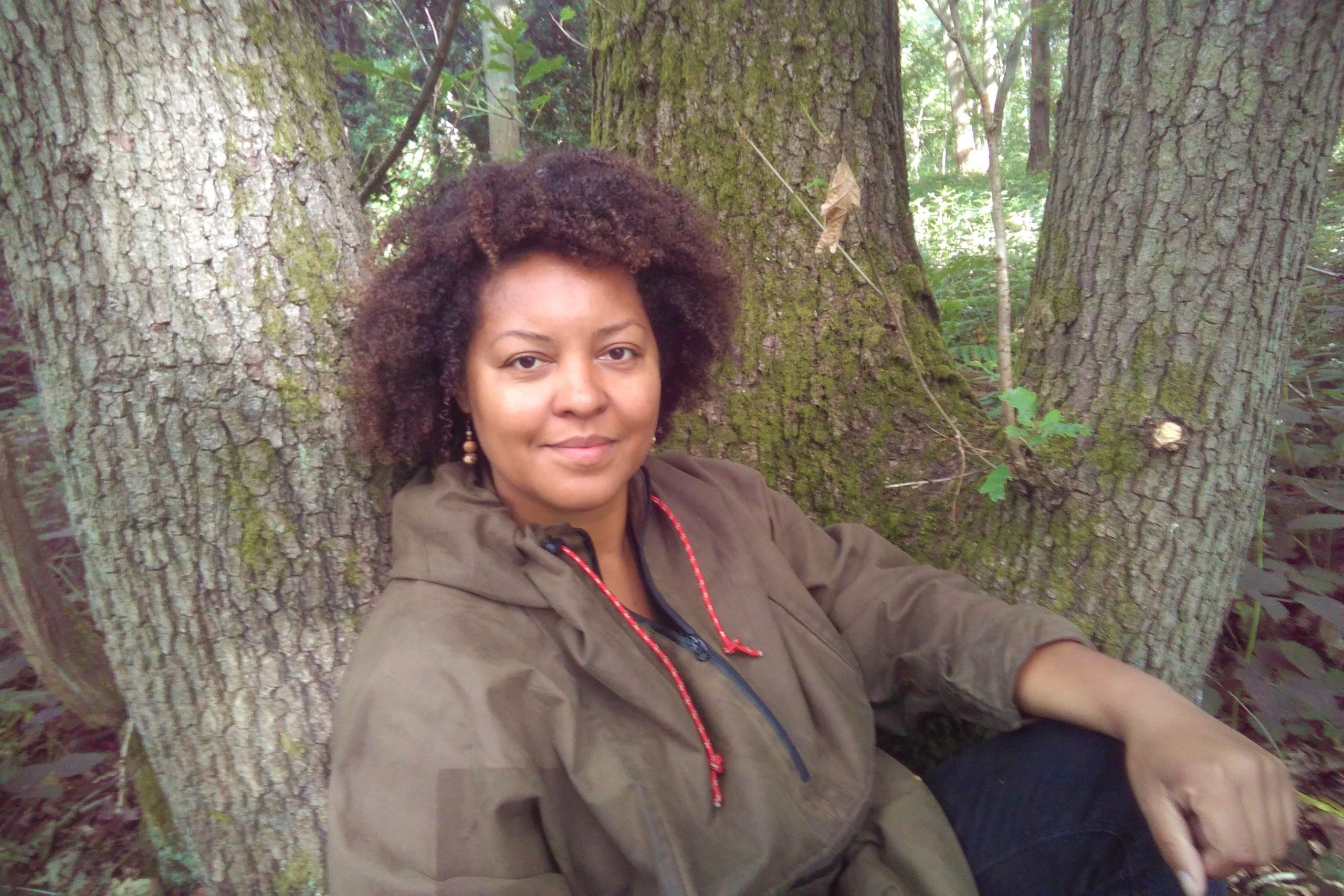 Beth sitting in front of a tree looking at the camera. She has tight curly brown hair and is wearing a dark green rain coat. Her arms is resting on her knee.