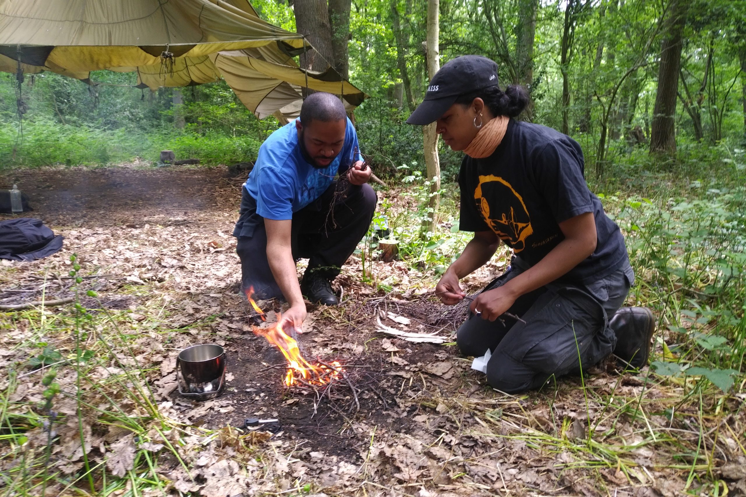 Two people building a camp fire in front of a tent in the woods. The man is wearing a blue t-shirt and dark waterproof trousers. The woman is wearing a black hat and t-shirt and waterproof trousers. They are kneeling on the ground.