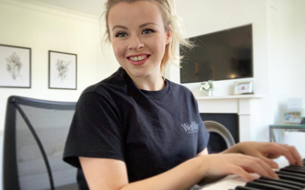 A blonde woman, Cate from Richmond performing arts school WestWay,, wearing a black t-shirt sitting playing a keyboard. Her blonde hair is tied up and she is turned to the camera smiling.