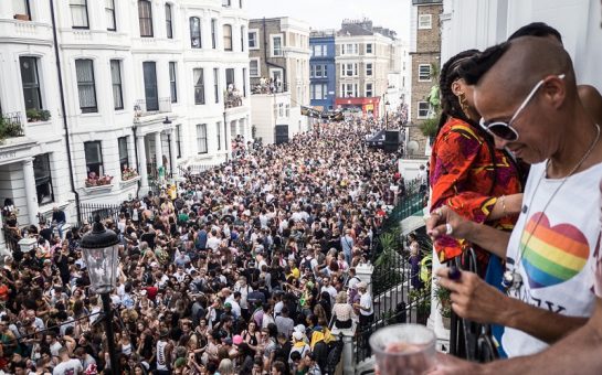 People looking over a balcony at a packed street of carnival goers. The person nearest the camera is wearing sunglasses and a t-shirt with a rainbow heart.