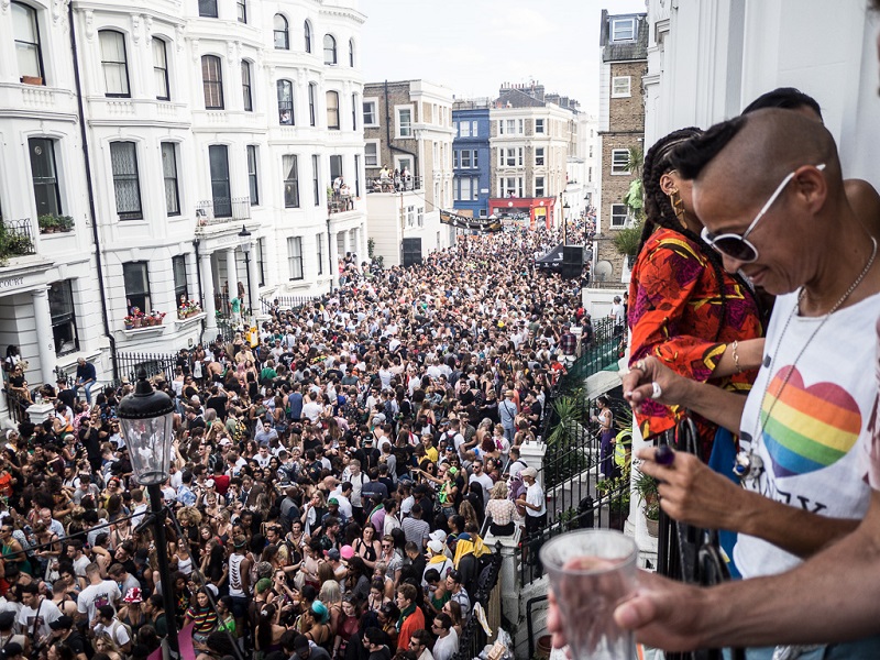 People looking over a balcony at a packed street of carnival goers. The person nearest the camera is wearing sunglasses and a t-shirt with a rainbow heart.