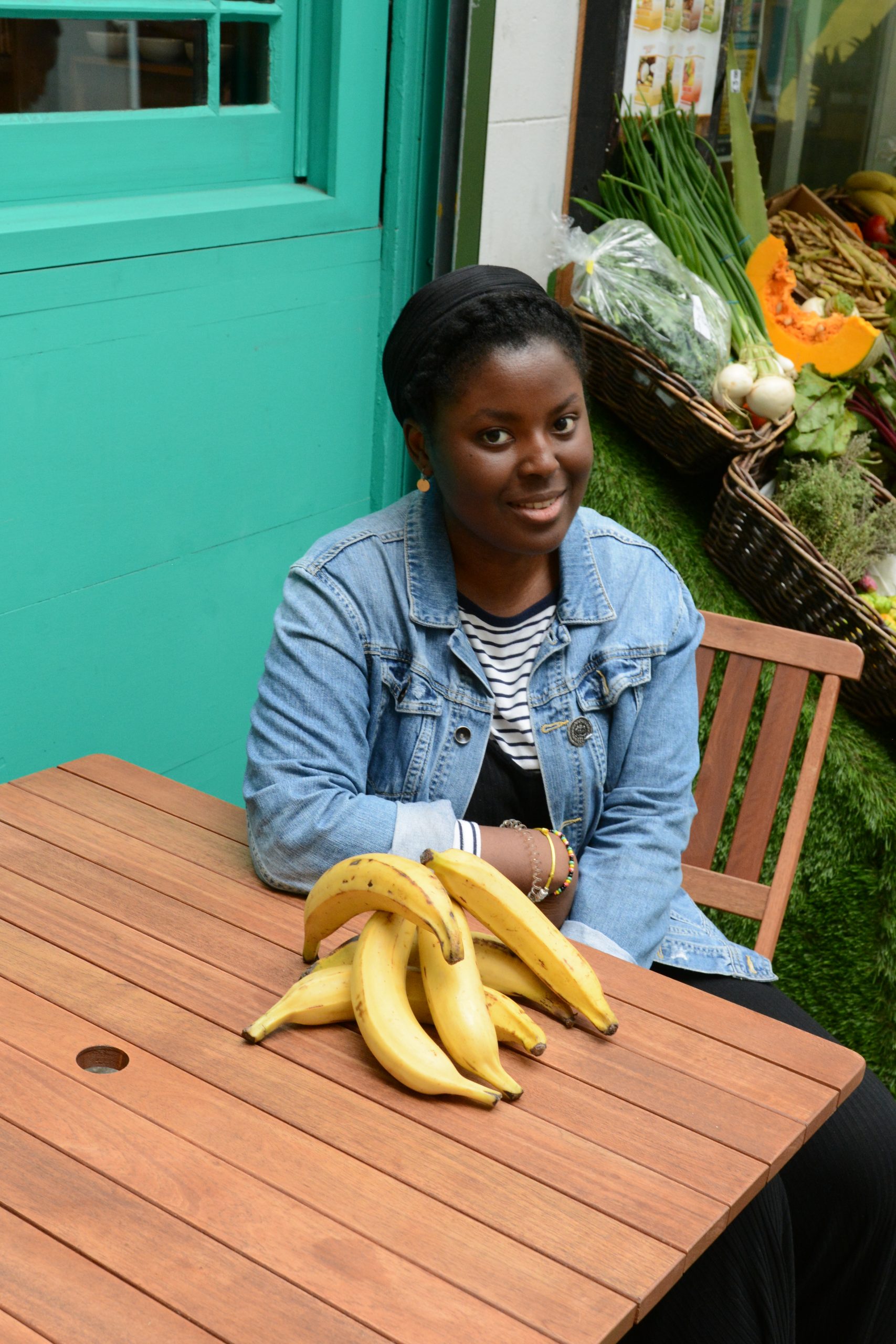 Tomi Makanjuola sits at a table. In front of her is a big bunch of plantains. Behind her there seems to be a fruit and veg stand.