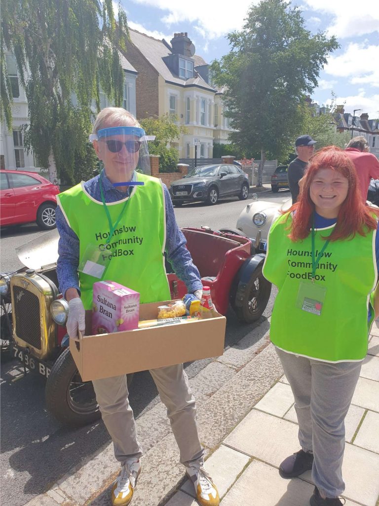 FoodBox volunteer Danielle with treasurer Philip Jones wearing high vis yellow vests. Philip is wearing a clear plastic visor over his face and is carrying a box full of supplied. They are standing in front of a red class Austin 7 car.