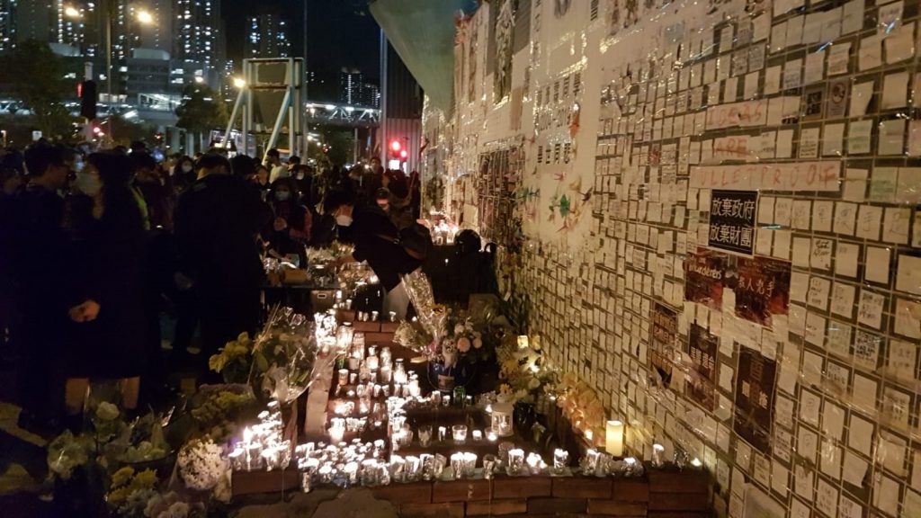 A crowd of people surrounding flowers and candles in the dark streets of Hong Kong.