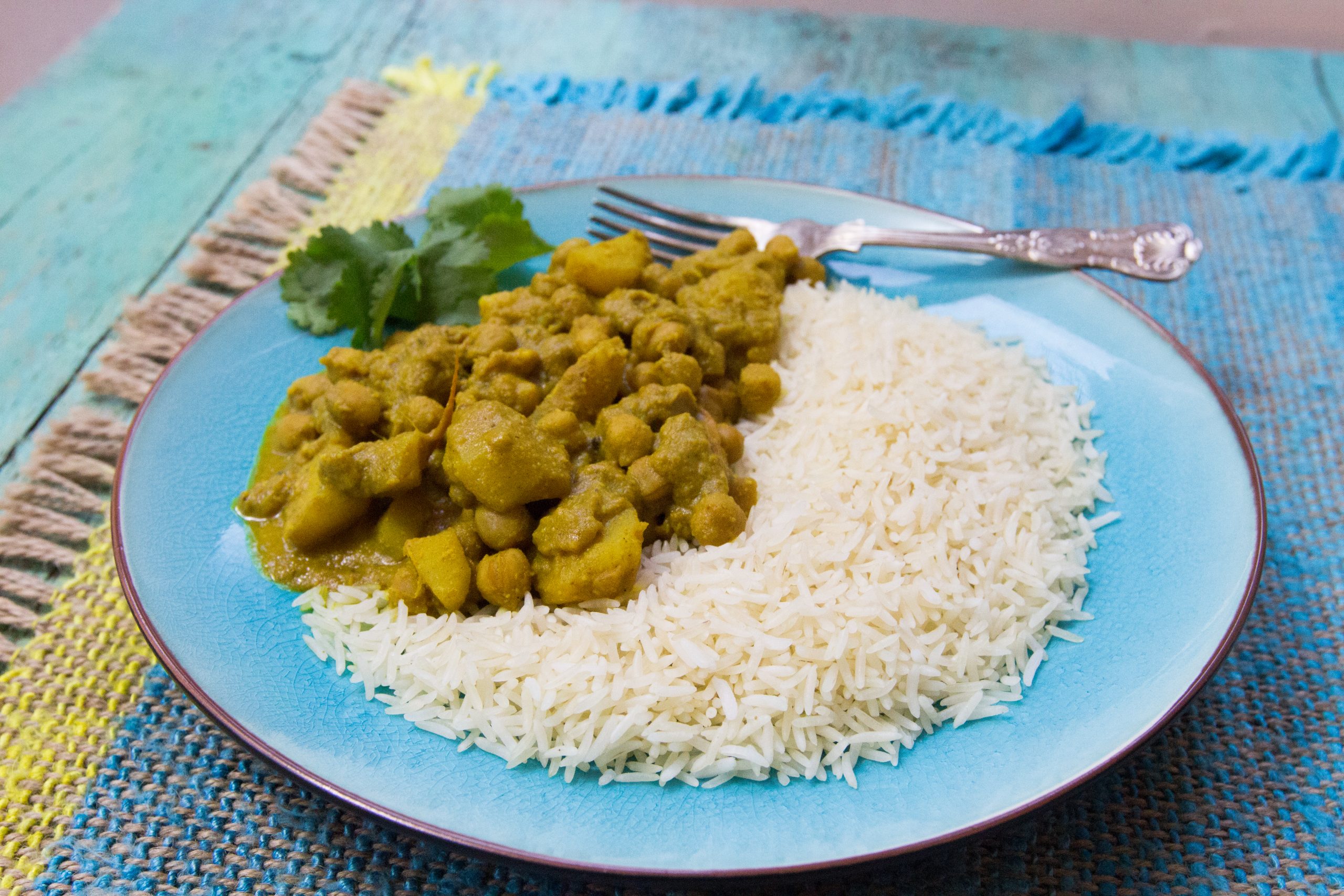 A chickpea curry with rice and a green garnish, set on a blue plate. There is an elegant silver fork sitting on the plate.