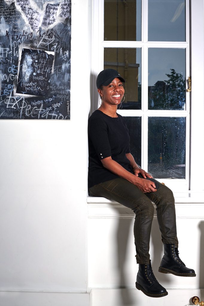 Linett Kamala, dressed in black, sitting in a white window, with her black and white artwork on the left. She is smiling.