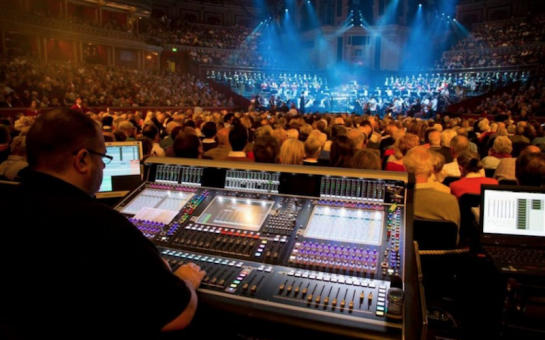 A DJ plays to a large crowd at an indoor music festival with lights in the background