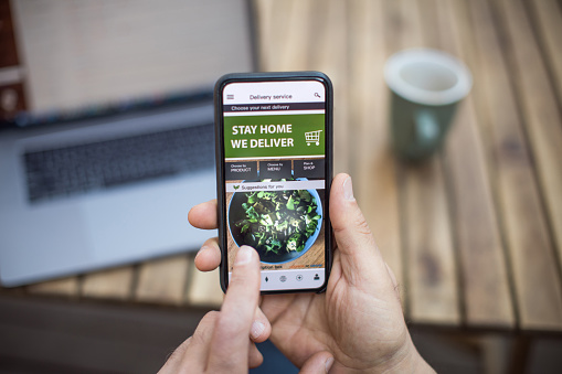 A stock image of a hand holding an iPhone doing a food shop online. You can see a blurred laptop and coffee cup on a wooden table in the background.