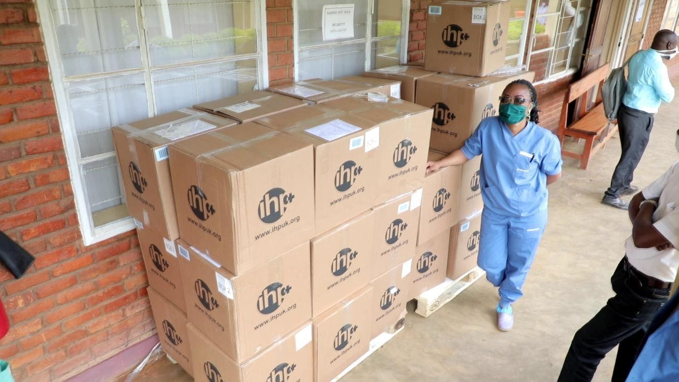 A woman in light blue scrubs wearing a dark screen face mask and sunglasses standing next to about 30 boxes piled taller than her.