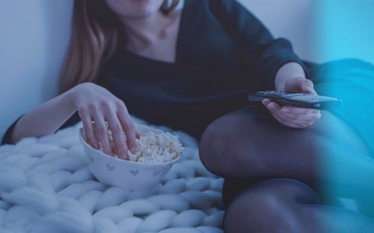 Photo of a woman reclining on a knitted blanket, with one hand in a bowl of popcorn and one hand holding a TV remote. You can't see her face, but you can see she's wearing red lipsticks, a dress and tights