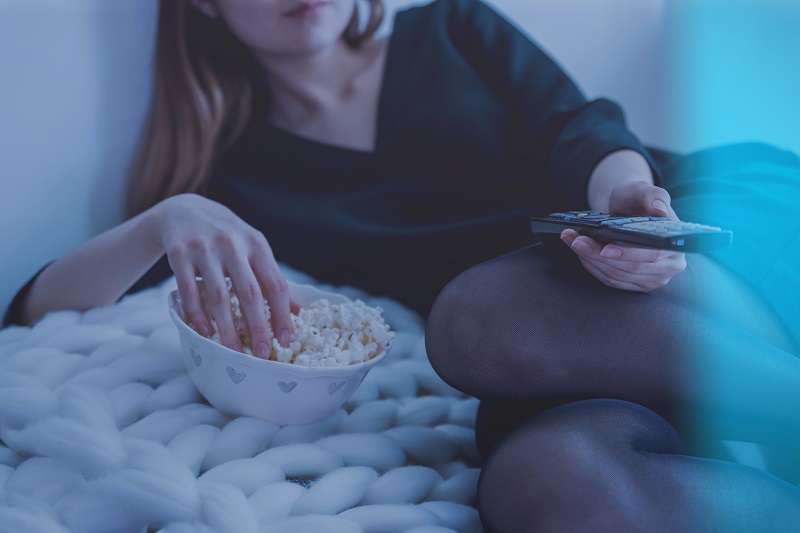 Photo of a woman reclining on a knitted blanket, with one hand in a bowl of popcorn and one hand holding a TV remote. You can't see her face, but you can see she's wearing red lipsticks, a dress and tights