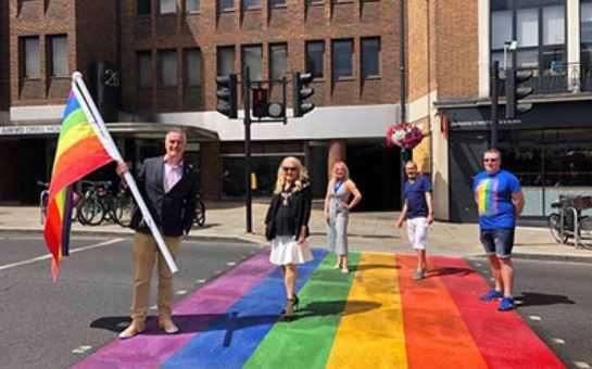 Five adults standing on the new Pride crossing in Richmond. The vertical lines stretch across the the whole road and are red, orange, yellow, green, blue and purple. One of the men is also carrying a large Pride flag. They all look happy and the sun is out.