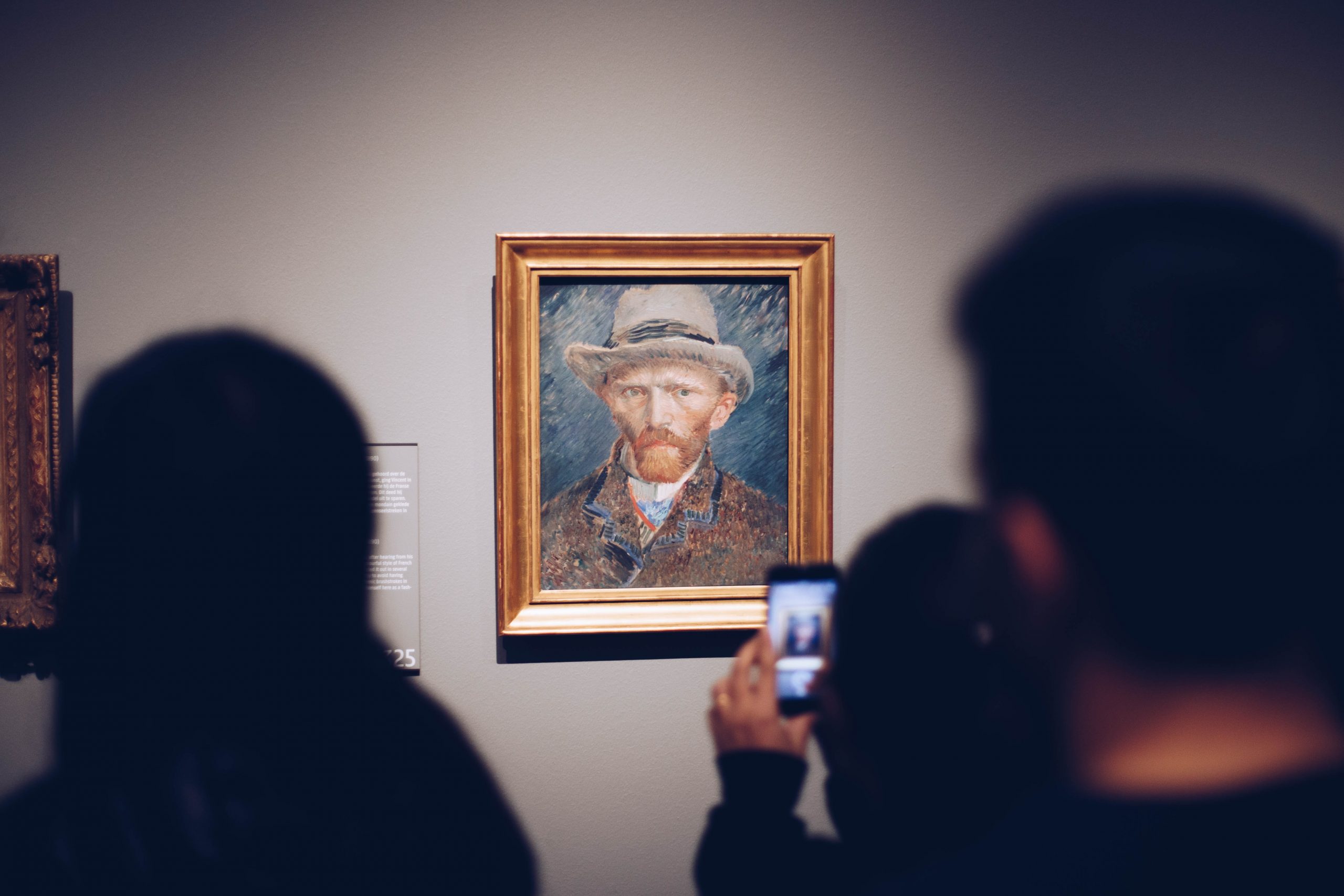 Image of a Van Gogh portrait on a wall, whilst in the foreground people are looking at it, one person taking a photo on their phone.