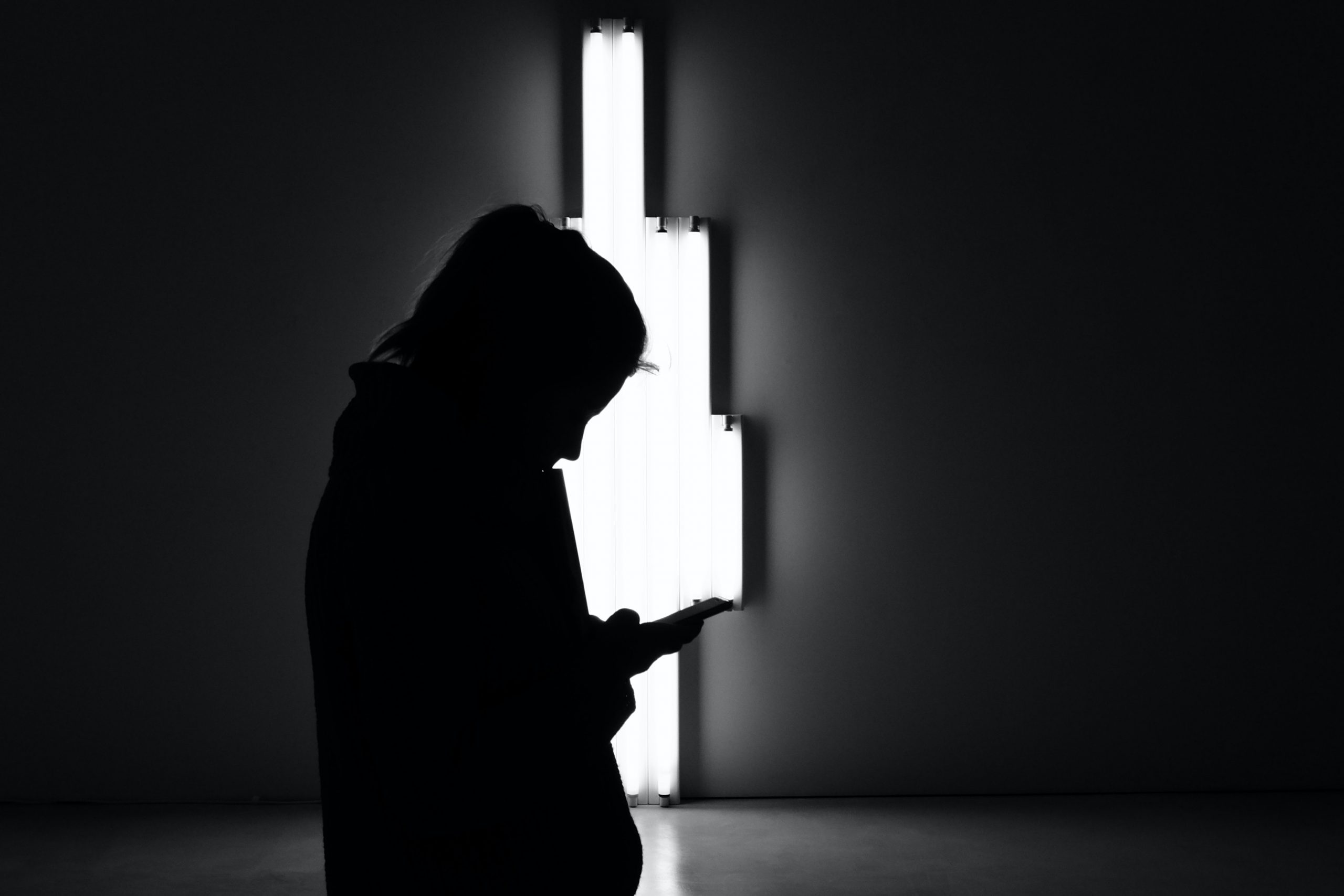 A silhouetted girl looks at her phone against a light installation. 