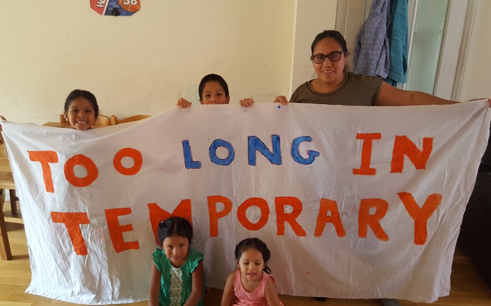 Janeth and her family hold a banner reading 'Too Long in Temporary'.