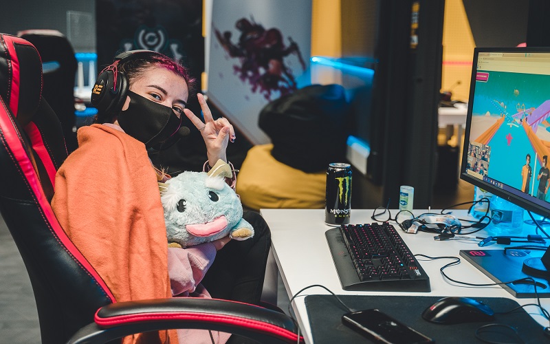 Jade Cavanagh gives a peace sign which wearing a face mask sat in front of a computer monitor in the esports arena