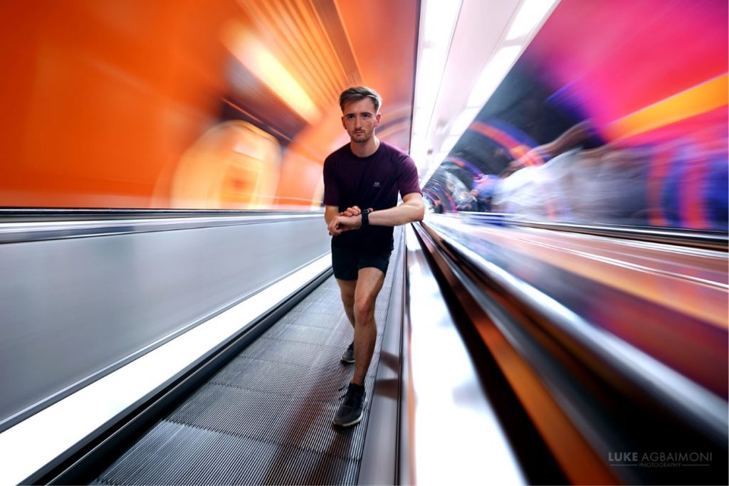 A blurry, busy red background. Matt "Runderground" Nelson poses for the camera on a tube station escalator.