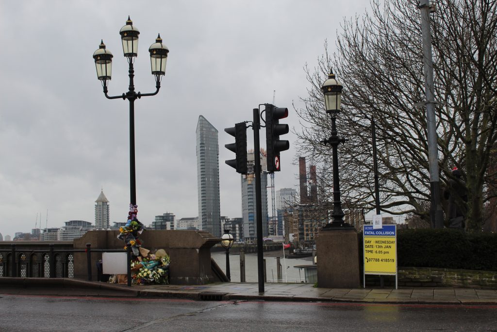 Picture of the site of the fatal collision on Battersea Bridge, showing flowers around a lamppost and a police sign calling for witnesses to the accident.