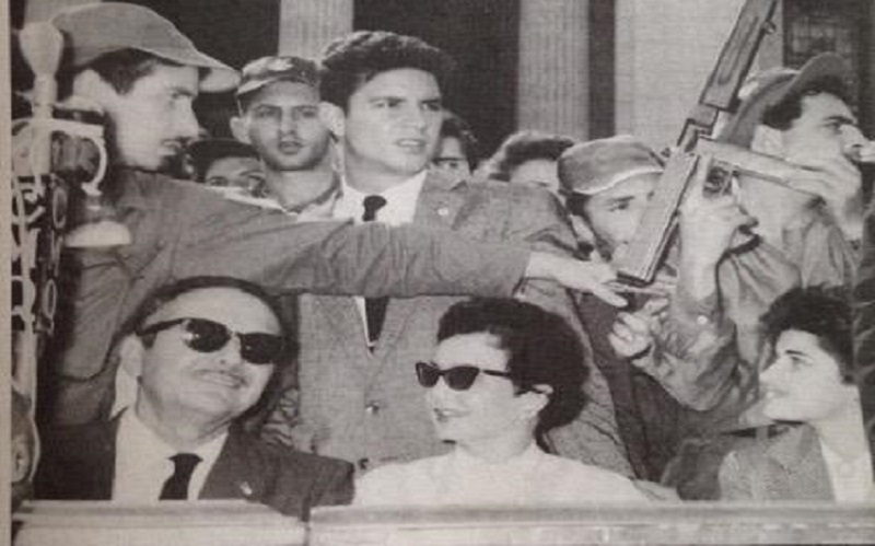 Picture of Helen's great aunt Ellie in Cuba, wearing sunglasses, in a crowd of people with a man holding a gun