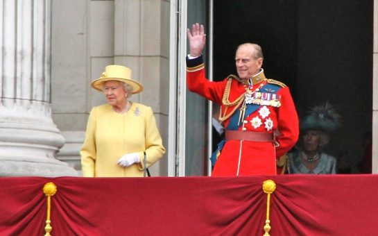 The queen in yelloe and prince philip in a military uniform waving from the balcony at buckingham palace