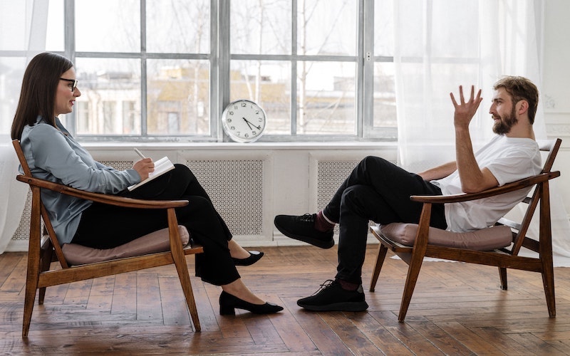 An image of a man and woman sitting across from each other as if she is taking notes like a drug and alcohol service therapist during Covid