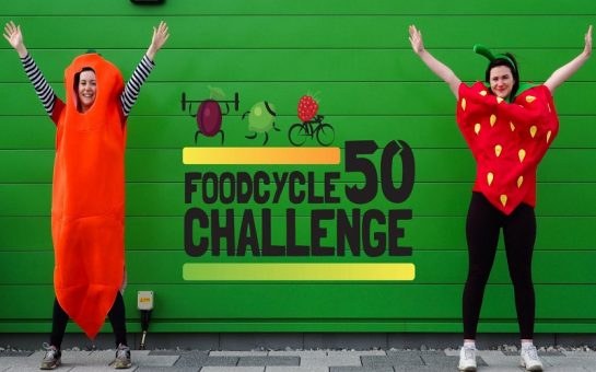 Two women, one dressed as a carrot, the other dressed as a strawberry, standing in front of a green wall with the words "FoodCycle 50 challenge" written on it.