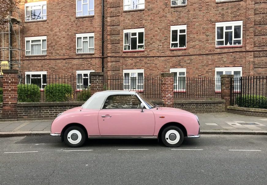A pink car parked on a road outside a block of flats.