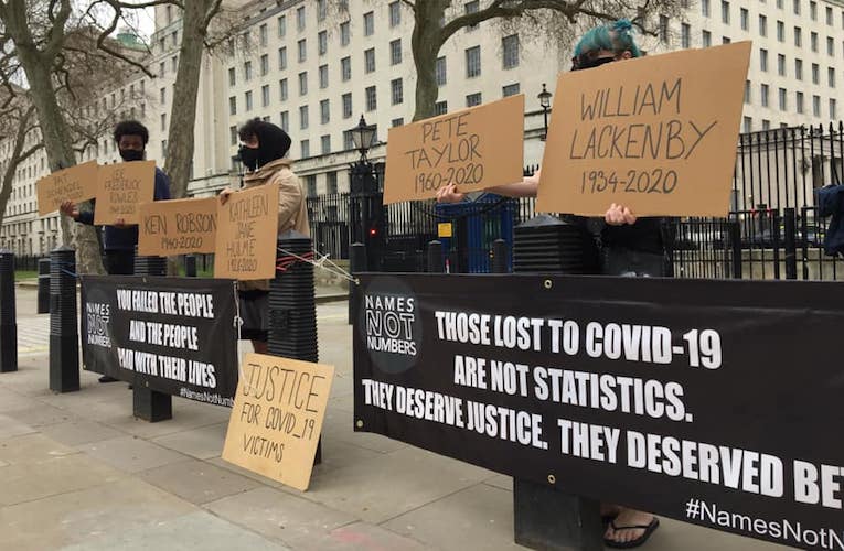 A group of campaigners are holding signs with the names of victims of COVID-19 in front of banners which read "You failed the people and the people paid with their lives" and "Those lost to COVID-19 are not statistics. They deserve justice. They deserved better."