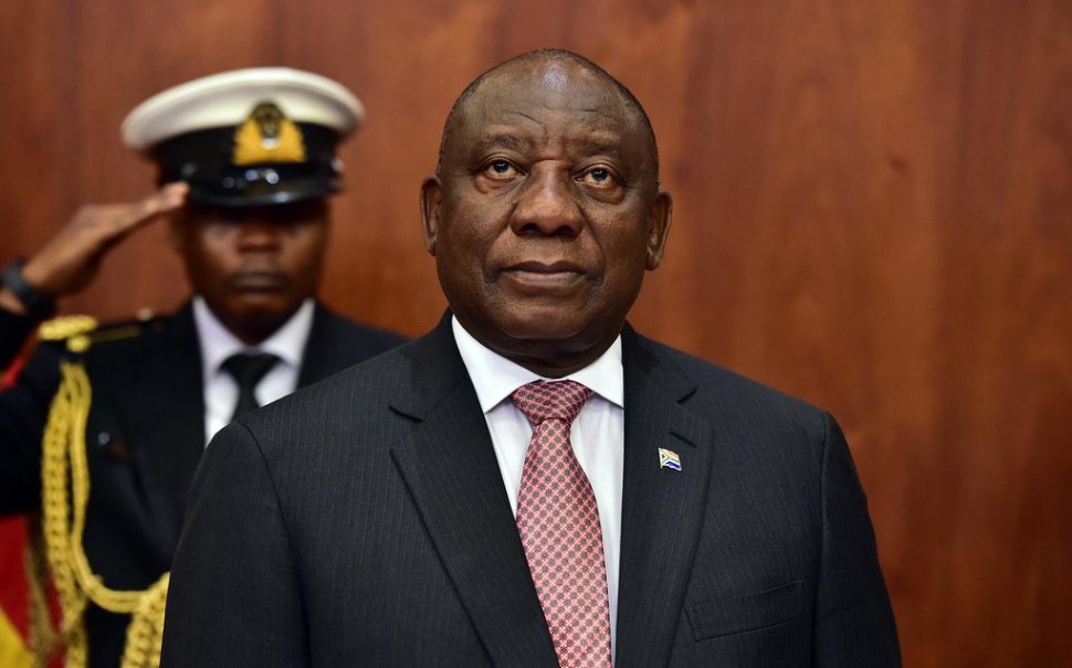President of South Africa, Cyril Ramaphosa stands in front of a saluting soldier with a brown background behind them.