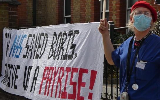 Sarah Jane Baker cheers in front of protest banner reading "THE NHS SAVED BORIS GIVE US A PAY RISE!". She is wearing scrubs, a face-mask, a stethoscope and a red beret outside Lewisham Hospital.