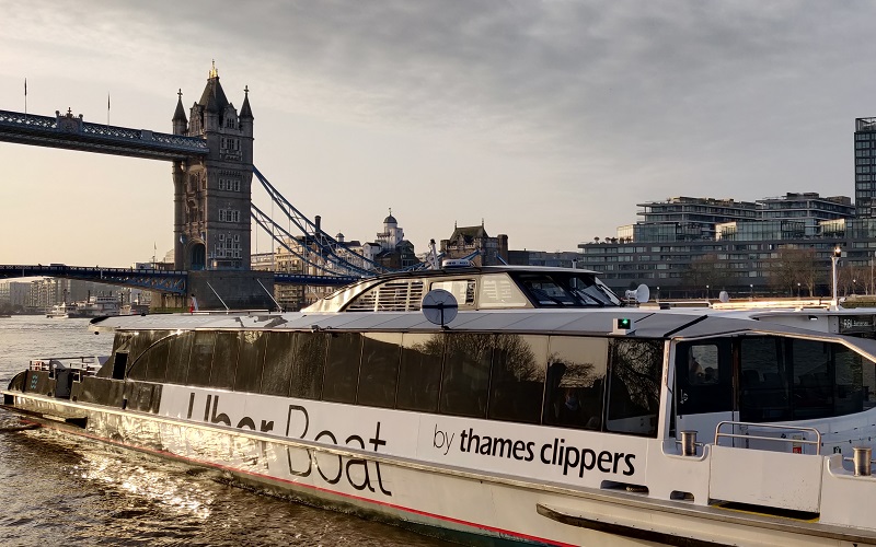 clipper boat approaches on a sunlit thames at dawn, tower bridge stands proud in the background with grey clouds overhead