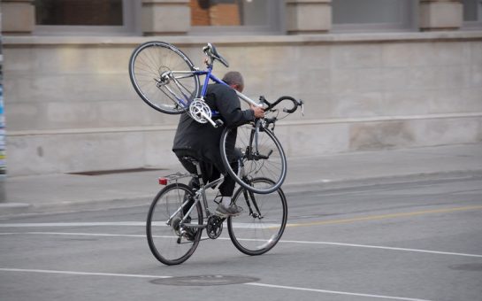 A man carries a bicycle over his shoulder as he rides away from the photographer on another bike