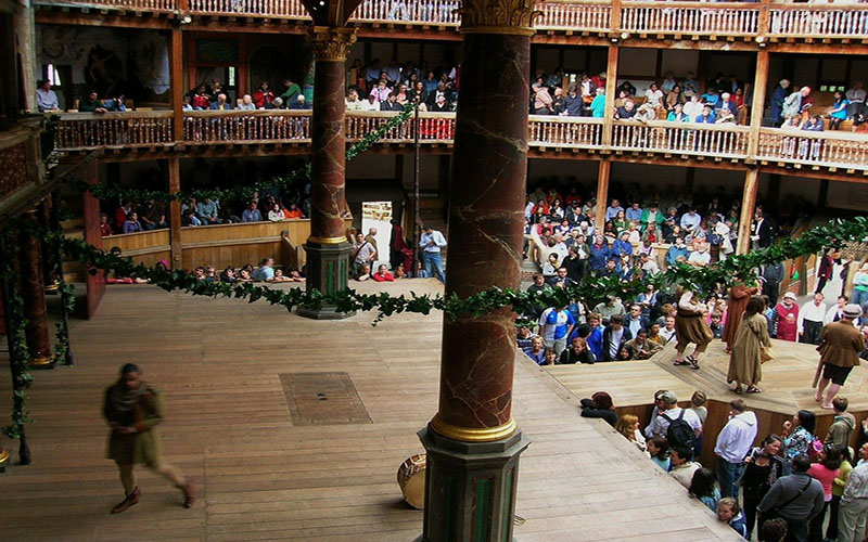 Inside of the Globe Theatre. The stage is wooden and theer are lots of wooden beams. The theatre is decorated with green leaf garlands. There are lots of people sitting in around the stage in tiered levels.