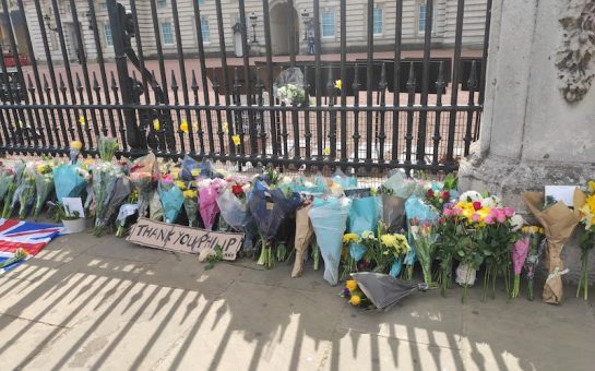 Flower bouquets lay outside Buckingham Palace. A cardboard sign reads "Thank you Philip"