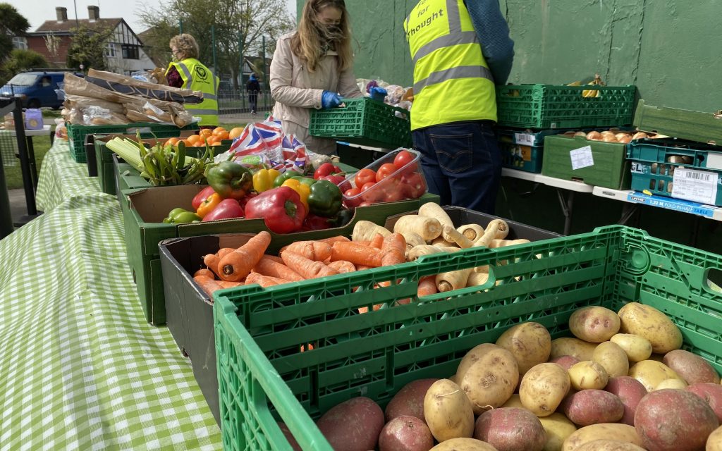 A long table with various boxes containing carrots, potatoes, peppers and other vegetables on display. Behind are 3 volunteers in green vests sorting the produce for customers.