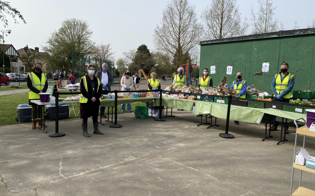 Wide angle shot showing the stall setup in its entirety. The 9 volunteers are standing behind the tables, posing for the photo. It is late afternoon and everyone is observing social distancing as well as wearing their masks and green vests for visibility.