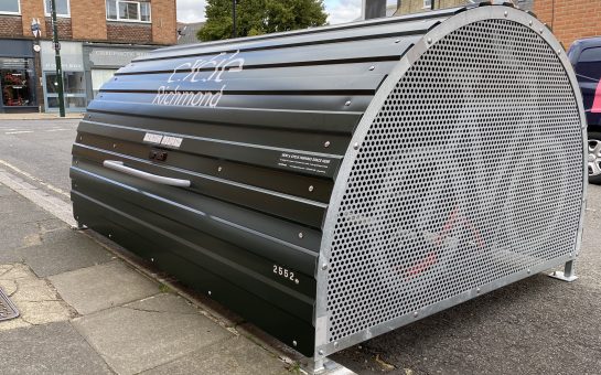 Side shot of the green half cylindrical bike hangar installed on a road. The hangar features an image of a bike on its side and has a metal lid in the centre.
