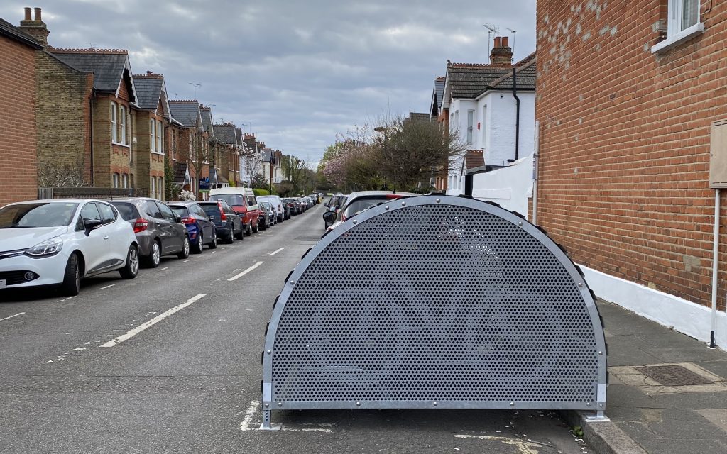 Side view of the bike hangar. It is placed on the right side of a street with cars lining the street on either side. It only takes a half of a lane up in size.