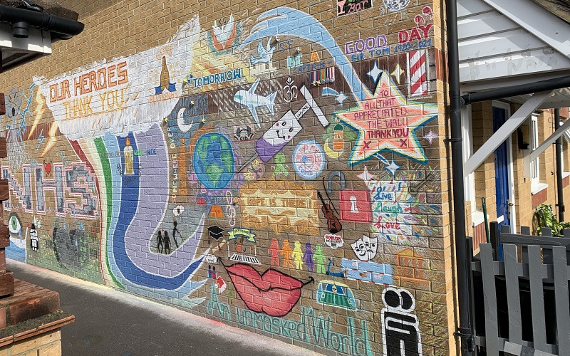 An image of the NHS mural featuring a pair of exposed lips captioned 'an unmasked world', a globe, a needle representing the vaccine, a football pitch, a white dove dedicated to Sarah Everard and a tribute to Captain Tom, amongst other details.
