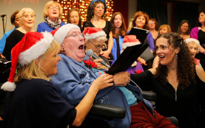 Sarah Cay holds the microphone to an old man who is singing in a chair while wearing a Christmas hat, Sarah is also singing. Choir of mixed ages sings behind him. A lady in a Christmas hat holds the music to him also singing.